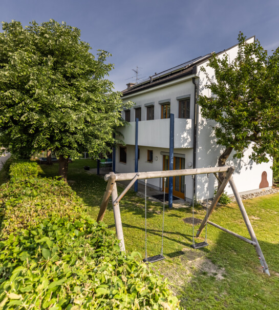 Außenansicht der Wohngemeinschaft mit Garten, Schaukel und abgrenzendem Gebüsch.