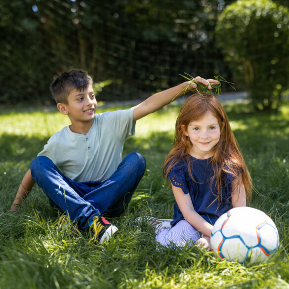 Zwei Kinder sitzen im Gras, das eine Kind legt dem anderen spielerisch Grashalme auf den Kopf.