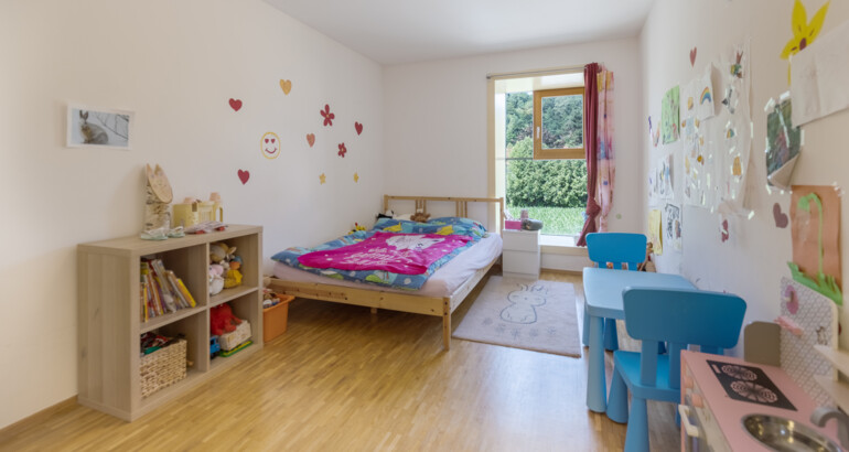 Innenansicht eines der Kinderzimmer der Wohngemeinschaft mit Bett, Spielküche, Regalen und Tisch mit Stühlen.