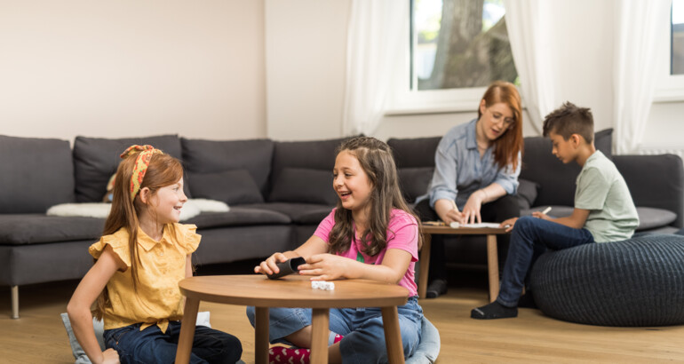 Zwei Kinder würfeln im Vordergrund, im Hintergrund zeichnet eine Sozialpädagogin mit einem Kind.