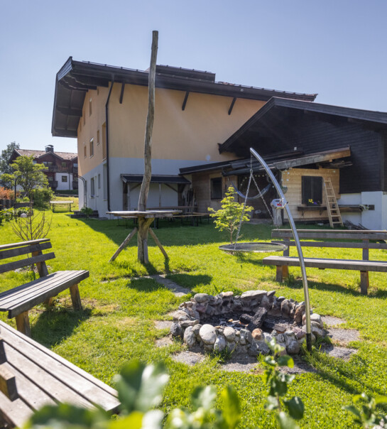 Außenansicht der Wohngemeinschaft mit Garten und Lagerfeuer-Stelle, umgeben von Bänken.